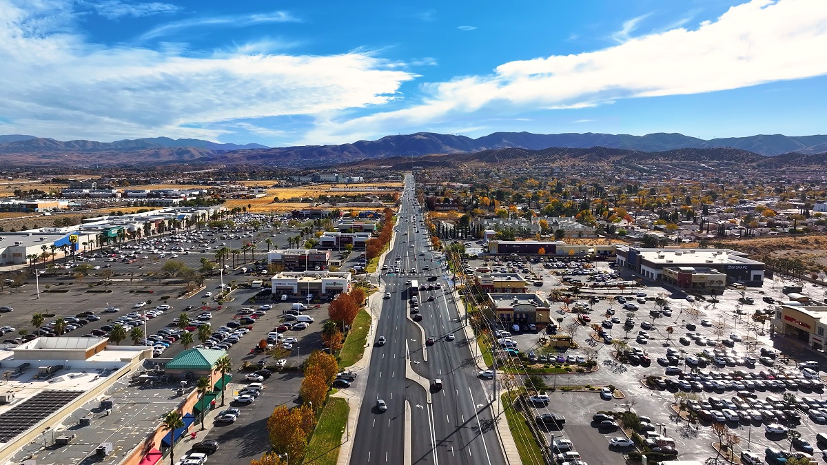 Aerial view of a bustling cityscape featuring a straight road flanked by parking lots, shops, and autumnal trees, with distant mountains under a bright blue sky.