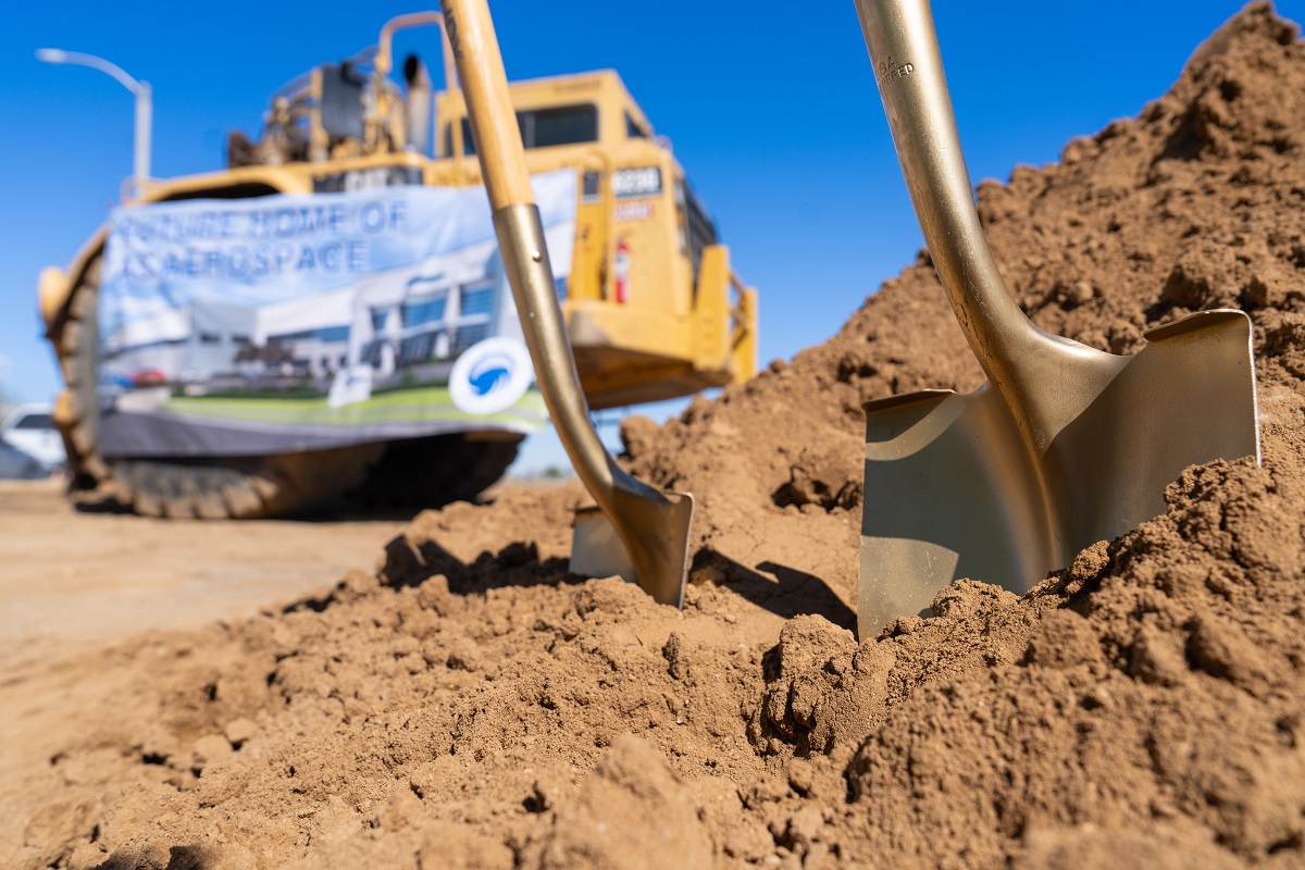 Golden shovels breaking ground in sandy soil, with a construction vehicle and a banner of an aerospace facility in the background under a clear blue sky.