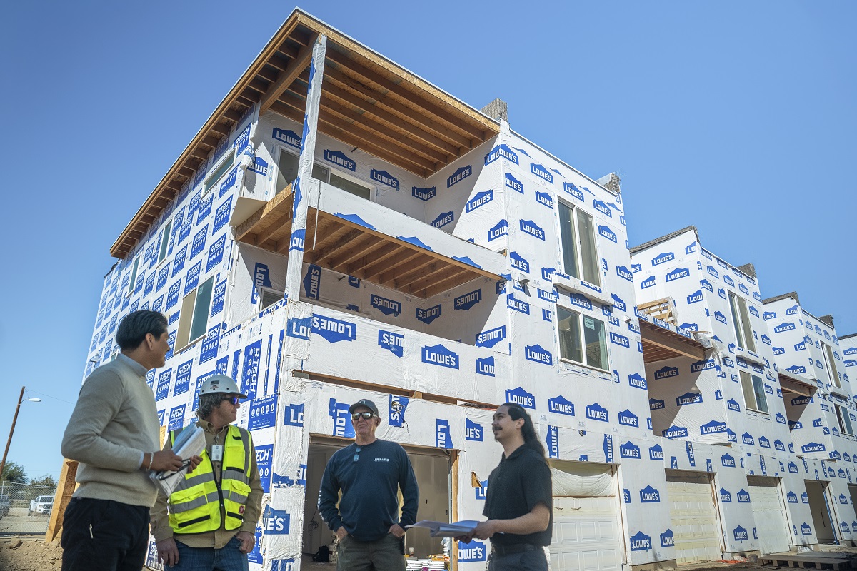 Four people in construction attire stand discussing in front of partially built townhouses covered in blue and white Lowe's wrap under a clear blue sky.