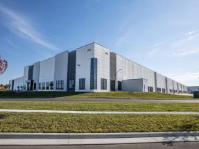 Large, modern industrial warehouse building with white and gray exterior, large windows, and a manicured lawn under a clear blue sky.