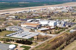 Aerial view of an industrial complex with a large parking lot filled with semi-truck trailers, office buildings, and an adjacent airport runway.