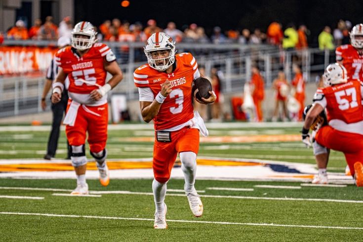 Football player in orange and white uniform runs with the ball on a field, teammates around.