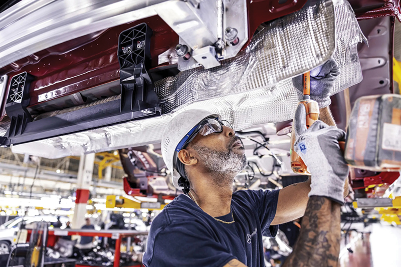 A factory worker wearing safety gear, including a white helmet and gloves, uses tools to assemble machinery overhead. Industrial setting, focused expression.