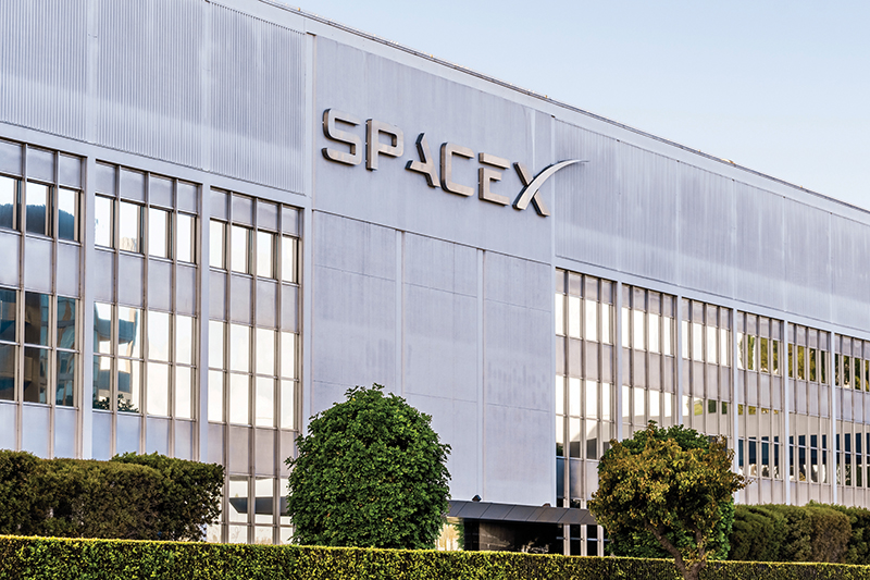 A modern building with the "SPACEX" logo prominently displayed above a wall of windows, surrounded by manicured hedges and trees.