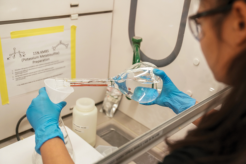 A person in blue gloves pours liquid from a flask into a funnel in a laboratory.