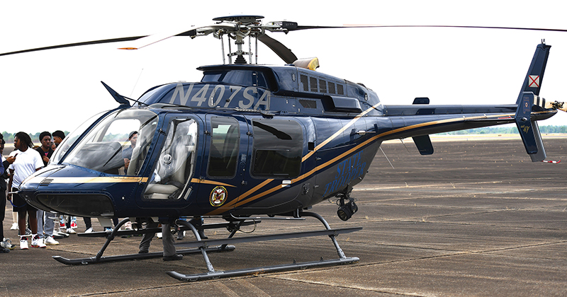 A dark blue Bell 407 helicopter with gold trim and registration N407SA is parked on a tarmac with people standing nearby.