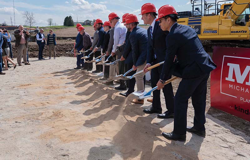 Groundbreaking ceremony with several people in hard hats using shovels to break ground near a Komatsu excavator and a sign.