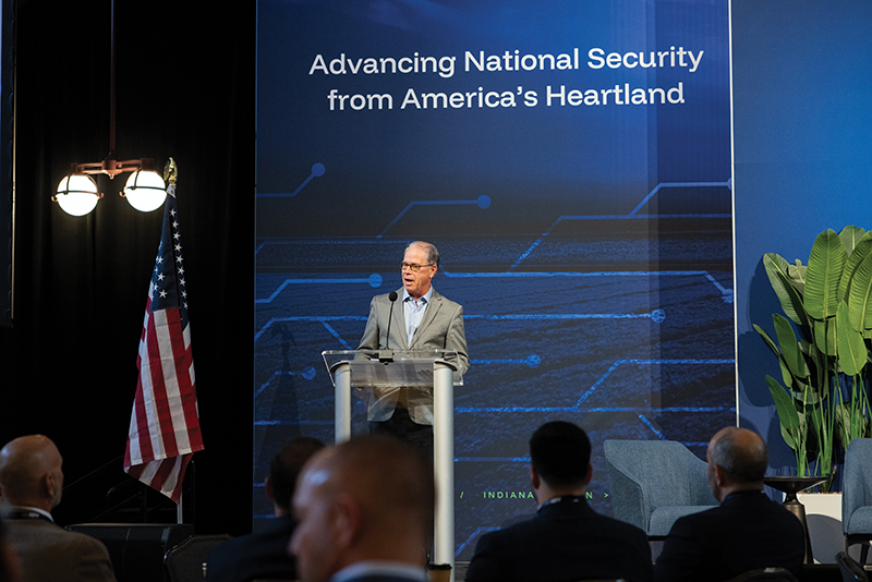 A man in a gray suit speaks at a podium in front of a banner that reads "Advancing National Security from America's Heartland."