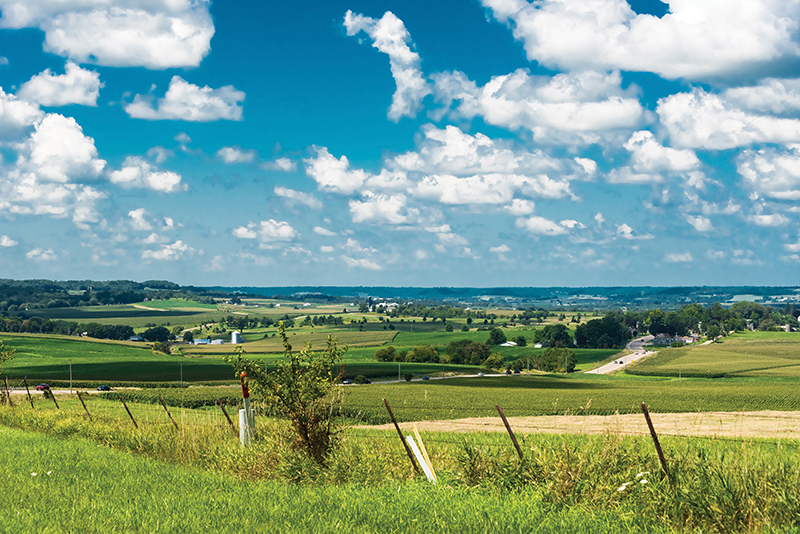 Rolling green hills and farmland under a bright blue sky with fluffy white clouds, featuring a distant road and farm buildings.