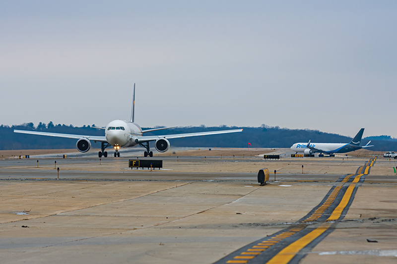 Two airplanes are on the tarmac, one a white passenger jet in the foreground and another a blue and white cargo plane in the background.